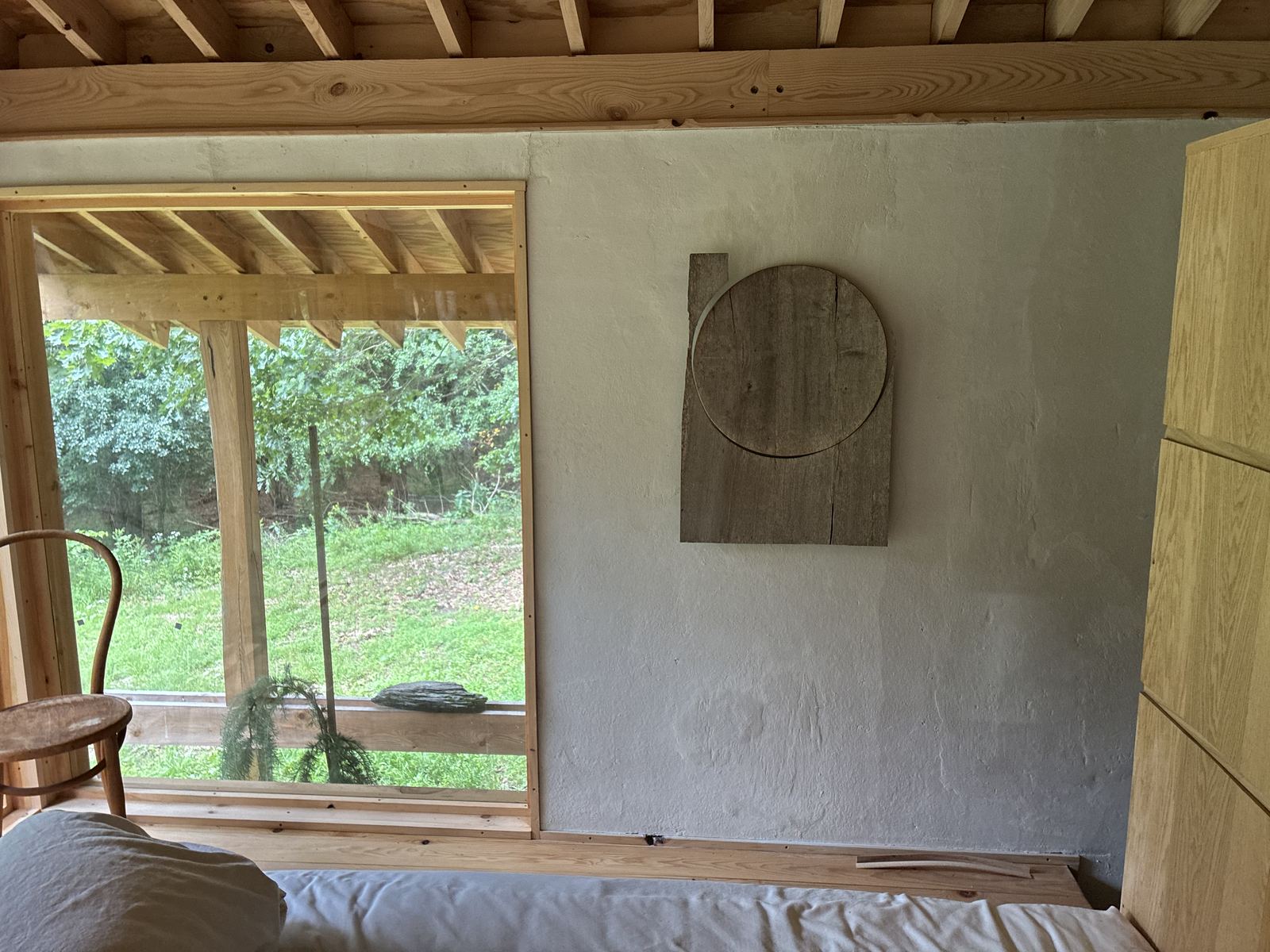Inside: a white plaster wall with a weathered wood relief, beside a large window framing green lawn and woods
